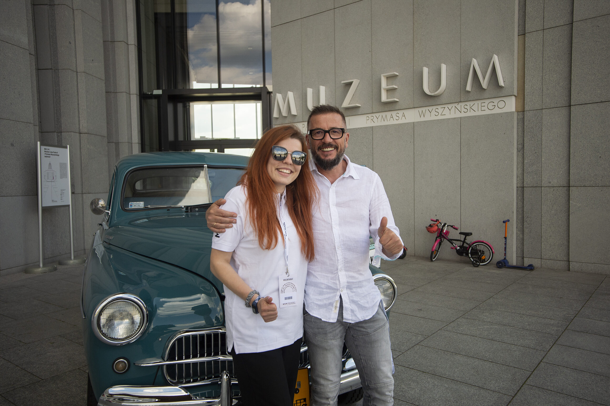 Slajd: Kobieta, mężczyzna w białych koszulach uśmiechnięci. Stoją przed samochodem, w tle na murze napis Muzeum.